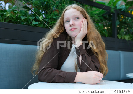 A beautiful red-haired girl, thoughtful, dreaming about something, sits at a table against a background of greenery. High quality photo 114163259
