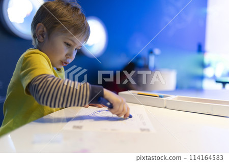 A young boy is drawing on a piece of paper with a blue crayon A young boy is drawing on a piece of paper with a blue crayon 114164583