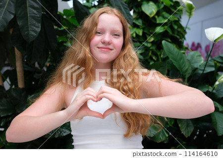 Beautiful red-haired, young woman, girl smiling, making a heart gesture against a background of greenery. High quality photo Beautiful red-haired, young woman, girl smiling, making a heart gesture against a background of greenery. High quality photo 114164610