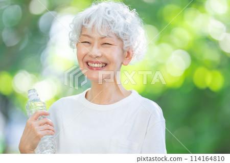 Senior woman drinking water in a park with fresh green leaves 114164810