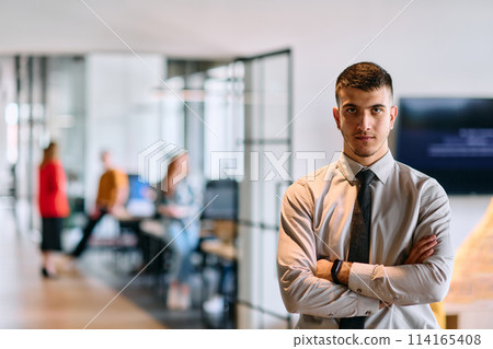 A young business leader stands with crossed arms in a modern office hallway, radiating confidence and a sense of purpose, embodying a dynamic and inspirational presence 114165408