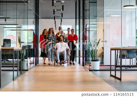 A diverse group of young business people walking a corridor in the glass-enclosed office of a modern startup, including a person in a wheelchair and a woman wearing a hijab 114165515