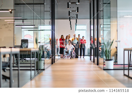 A diverse group of young business people walking a corridor in the glass-enclosed office of a modern startup, including a person in a wheelchair and a woman wearing a hijab 114165518