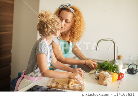 Beautiful young mom and her cute little daughter rolling out dough at home. 114165621
