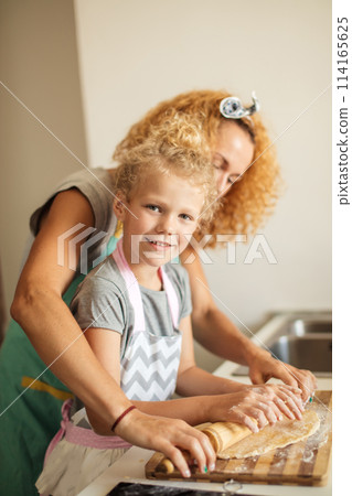 Beautiful young mom and her cute little daughter rolling out dough at home. 114165625