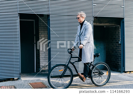 Stylish male in checked coat and sunglasses riding on retro bicycle near gray urban wall in sunny day. Neutral carbon footprint transportation. Green eco friendly mobility sustainable transport 114166293