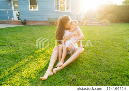 Cheerful Caucasian preschooler kid is embraced by happy young mother, looking at camera, smiling. Young mom and kid hugging with love, affection, tenderness on the backyard lawn background on sunset. 114166314