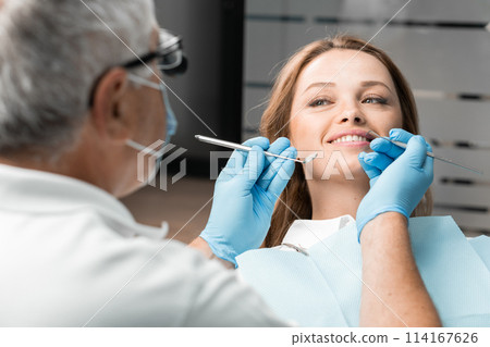 A patient in a dental chair happily reveals her beautiful smile made thanks to the treatment dentist. Doctor who performed procedure is happy with the look it and strives to provide long-term support. 114167626