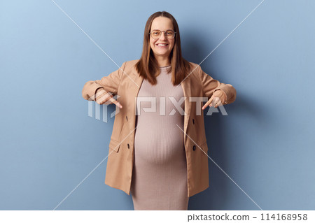 Positive smiling indicating pregnant woman in beautiful dress and jacket posing isolated over blue background looking at camera with satisfied face indicating at her belly 114168958