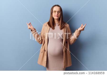 Calm relaxed pregnant elegant woman in beautiful dress and jacket posing isolated over blue background standing with mudra gesture meditating trying to calm down 114168959