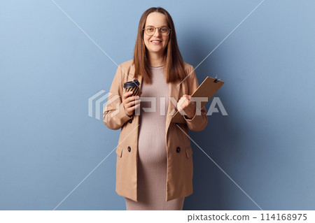 Smiling cheerful pregnant businesswoman in beautiful dress and jacket posing isolated over blue background holding clipboard with documents and enjoying takeaway coffee 114168975