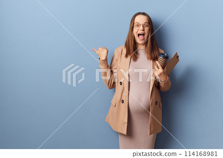 Excited pregnant woman in beautiful dress and jacket posing isolated over blue background showing advertisement area pointing at copy space for advertisement exclaiming happily 114168981