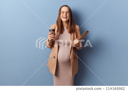 Laughing pregnant businesswoman in beautiful dress and jacket posing isolated over blue background having break from work drinking hot coffee expressing happiness Laughing pregnant businesswoman in beautiful dress and jacket posing isolated over blue background having break from work drinking hot coffee expressing happiness 114168982