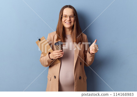Cheerful pregnant woman in beautiful dress and jacket posing isolated over blue background showing like gesture thumb up enjoying her work holding coffee and clipboard 114168983