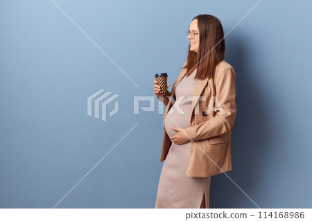 Side view of pregnant woman in beautiful dress and jacket posing isolated over blue background looking away with takeaway coffee in hands copy space for advertisement 114168986