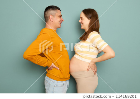 Funny cheerful family waiting baby isolated over blue background standing with hands on hips measuring bellies expectant parents having fun during pregnancy 114169201