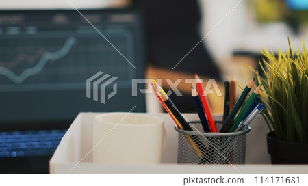 Focus on desk belongings in modern office with notebook in blurry background showing business diagram and figures, close up. Pencil holder, mini house plant, empty coffee mug and graphs on laptop Focus on desk belongings in modern office with notebook in blurry background showing business diagram and figures, close up. Pencil holder, mini house plant, empty coffee mug and graphs on laptop 114171681