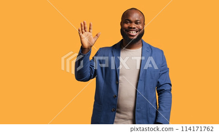African american man cheerfully doing salutation hand gesture. Portrait of happy BIPOC person raising arm to greet someone, gesturing, isolated over yellow studio background, camera A African american man cheerfully doing salutation hand gesture. Portrait of happy BIPOC person raising arm to greet someone, gesturing, isolated over yellow studio background, camera A 114171762