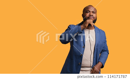 Upbeat african american singer holding microphone, performing tunes, isolated over yellow studio background. Energetic musician singing to imaginary crowd, doing karaoke, camera B Upbeat african american singer holding microphone, performing tunes, isolated over yellow studio background. Energetic musician singing to imaginary crowd, doing karaoke, camera B 114171787