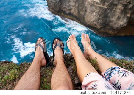 A couple sits on a cliff with their feet dangling over an abyss above the ocean. First person photo. A couple sits on a cliff with their feet dangling over an abyss above the ocean. First person photo. 114171824