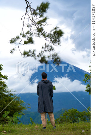 A young man stands with his back in a black raincoat enjoying the view of the popular sacred mountain Volcano Agung, shrouded in clouds on the island of Bali. A young man stands with his back in a black raincoat enjoying the view of the popular sacred mountain Volcano Agung, shrouded in clouds on the island of Bali. 114171857