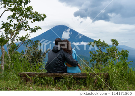 A couple of lovers are sitting on a bench, hugging each other and enjoying the view of the popular sacred cloud-covered Mount Agung on the island of Bali. A couple of lovers are sitting on a bench, hugging each other and enjoying the view of the popular sacred cloud-covered Mount Agung on the island of Bali. 114171860