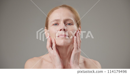 Real people age, beauty, health and dry skin care concept - beautiful mature Caucasian middle aged woman in her 50s touching facial skin doing facial massage and looking at camera 114179496