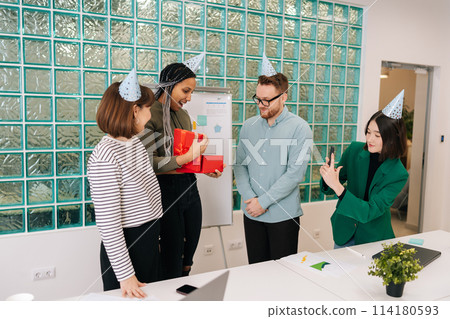 Friendly team of multiethnic business people congratulate African-American female colleague and give gift with surprise. Cheerful birthday woman surprised to open box with gift by around coworkers. 114180593