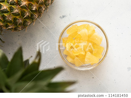 Dried soft sweet pineapple slices in glass bowl with raw pineapple on light table. 114181450