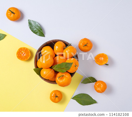 sweet and juicy tangerines with green leaves in a wooden bowl on a trendy yellow-gray background. sweet and juicy tangerines with green leaves in a wooden bowl on a trendy yellow-gray background. 114182108