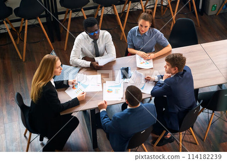 Female CEO leads diverse team in strategy session at modern office, with charts, laptop for data-driven decision making. Professionals engage in creative solutions, collaboration for business growth. 114182239