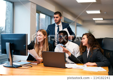Multiethnic team works in modern office, young woman points at computer screen, presenting ideas. Colleagues gather around desk with laptop, paperwork, engaging in creative group discussion. 114182241