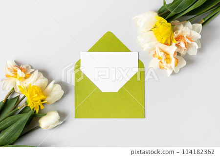 Green envelope with clean white sheet of paper for your text and spring flowers white tulips and daffodils on gray background. Invitation to the holidays. Flat lay. 114182362