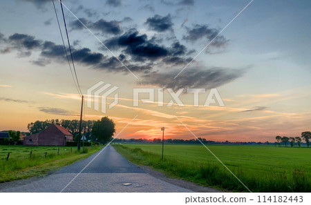Dramatic Sunrise Over Rural Road with Farmhouse and Lush Fields 114182443