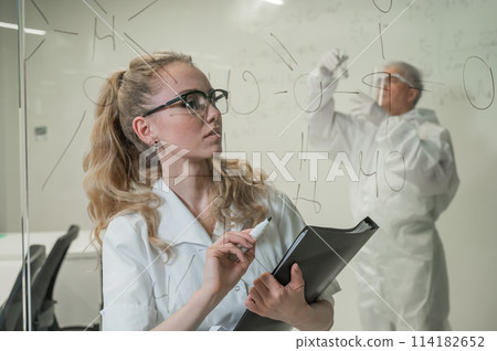 A woman chemist writes a formula on glass. An elderly Caucasian man in a protective suit is doing tests. 114182652