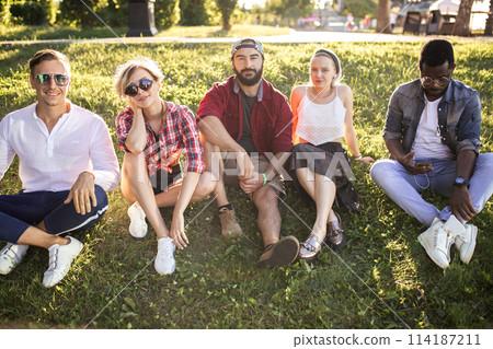 Diverse group of cheerful friends sitting on grass at sunset in nature park. Diverse group of cheerful friends sitting on grass at sunset in nature park. 114187211