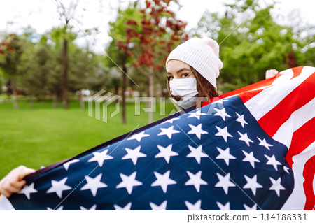 Girl in protective sterile medical mask and with american flag in hand. Quarantine city. Covid -19. Girl in protective sterile medical mask and with american flag in hand. Quarantine city. Covid -19. 114188331