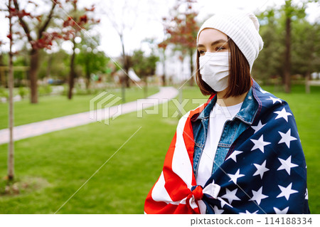 Girl in protective sterile medical mask and with american flag in hand. Quarantine city. Covid -19. Girl in protective sterile medical mask and with american flag in hand. Quarantine city. Covid -19. 114188334