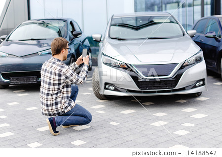 Man customer taking photo to mobile phone chooses auto and wants to buy new car in showroom Man customer taking photo to mobile phone chooses auto and wants to buy new car in showroom 114188542