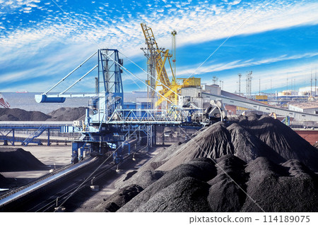 Coal harbor at maritime dry bulk cargo terminal, bucket wheel stacker reclaimer reloads coal onto an automatic conveyor belt. Coal harbor at maritime dry bulk cargo terminal, bucket wheel stacker reclaimer reloads coal onto an automatic conveyor belt. 114189075