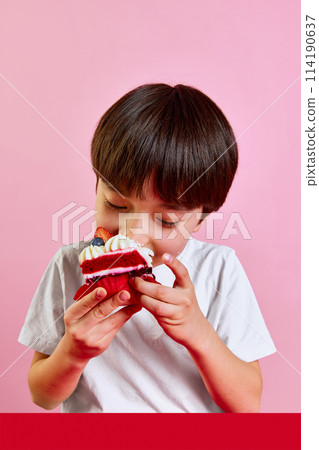 Delightful dessert time. Boy, kid enjoying delicious red velvet cake with cream and berries, making big bite against pink background Delightful dessert time. Boy, kid enjoying delicious red velvet cake with cream and berries, making big bite against pink background 114190637