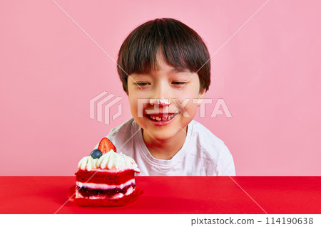 Little boy eating red velvet cake decorated with strawberries, with dirty face of cream against pink background Little boy eating red velvet cake decorated with strawberries, with dirty face of cream against pink background 114190638