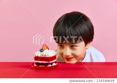 Happy little Korean boy leaning head on table and looking at delicious cake with intense to eat against pink background Happy little Korean boy leaning head on table and looking at delicious cake with intense to eat against pink background 114190639