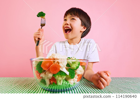 Playful and healthy. Smiling boy, kid raising for with broccoli, sitting at table with bowl full of boiled vegetables against pink background Playful and healthy. Smiling boy, kid raising for with broccoli, sitting at table with bowl full of boiled vegetables against pink background 114190640
