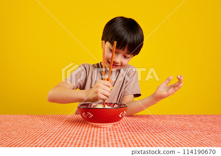 Smiling boy sitting at table and with plate of ramen soup and learning to eat with chopsticks against yellow background 114190670