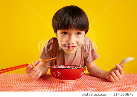 Cheerful young boy using chopsticks and spoon, sitting at table and eating ramen, noodle soup against yellow background Cheerful young boy using chopsticks and spoon, sitting at table and eating ramen, noodle soup against yellow background 114190672