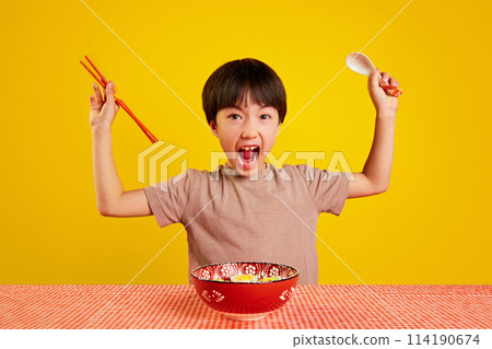 Little Korean boy, child holding chopsticks and spoon, emotionally sitting at table with ramen soup against yellow background Little Korean boy, child holding chopsticks and spoon, emotionally sitting at table with ramen soup against yellow background 114190674