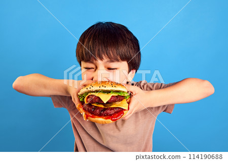 Moment of pleasure. Little boy, kid biting delicious giant burger with meat, vegetables and cheese against blue background 114190688