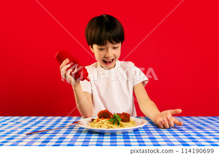 Playful child, little boy sitting at table and squeezing ketchup sauce on plate with pasta and meatballs against red background 114190699