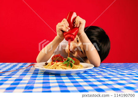 Playful kid, little boy sitting at table and squeezing ketchup sauce on plate with spaghetti and meatballs against red background 114190700